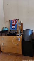 Wide shot of wooden dresser with resin waste bin, metal storage boxes, globe, cork board, desk lamp, flags, and pencils on and around it