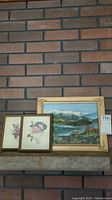 Photo showing the three framed artworks set against a brick wall: a large gold-tone framed landscape painting and two smaller framed rose prints.