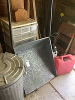Galvanized metal garbage can with domed lid and galvanized metal snow pusher with handle along with red plastic jerry can and white garden sprayer