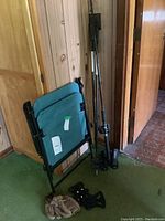 Folding lawn chair next to fishing rod, ski poles, brown baseball glove, and shoe covers on green carpeted floor against wooden wall.