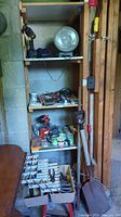 Shelving unit holding various hardware supplies including paint cans, drill, work light, and shovel leaning against wall.