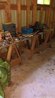 Photo showing three wooden saw horses with a long piece of plywood on top serving as a workbench, with various tools and items on the surface. Barn interior with wood framing and concrete floor visible.
