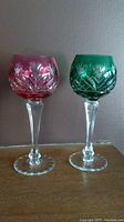 Pair of vintage Bohemian crystal hock wine glasses, one emerald green and one amethyst purple, placed side-by-side on a wooden surface against a plain wall.