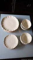 Overview of dinner plates, lunch plates and bowls stacked separately showing scalloped edges and olive branch detail on light beige stoneware.