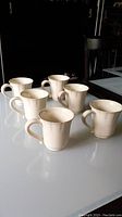 Group of 6 cream-colored Costa Nova stoneware mugs arranged on a white reflective surface, showing their shape and handles.