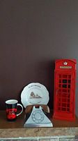 Photo showing the full lot on a shelf: black and red telephone-themed mug, white decorative plate with Bell Homestead image, silver metal rotary phone with wood handset, and tall red British telephone booth decorative piece.