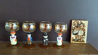 Four Goebel Hummel crystal wine glasses displayed in a row with ceramic figurine stems and gold etched bowls next to a framed Hummel print.