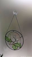 View of the hanging stained glass bird suncatcher, showing white bird with green leaves, circular black frame, and hanging chain attached to window suction hook.