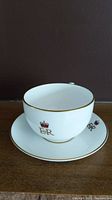 White fine bone china teacup and saucer set photographed against a dark background on a wooden surface. Both pieces decorated with gold trim and Queen Elizabeth II insignia.