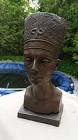 Front side of bronze-tone Queen Nefertiti bust statue with tall textured crown and square base, outdoors background.