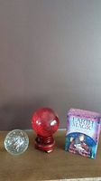 Three items: red glass crystal ball on wooden stand, clear glass paperweight with bubbles and swirl, Earth Magic Oracle card deck in purple box, all displayed on wooden surface.