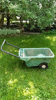 Side view of Scotts garden cart showing green plastic tub, metal handles, and wheel setup on grass near garden.