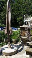 Folded beige Sun Garden Easy Sun Parasol umbrella attached to heavy round concrete base placed on outdoor stone patio surrounded by plants and garden decor.
