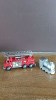 Miniature Timex firetruck clock in red metal alongside a silver Chass train clock, both heavy for their size.