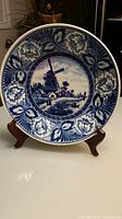 Full view of blue and white Delft plate displayed on wooden stand showing windmill in center and floral rim.