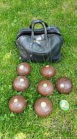 Seven wooden lawn bowling balls arranged outdoors on grass with vintage black leather bag behind.