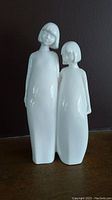Full frontal view of two white porcelain sister figurines side by side on a flat surface with dark background.