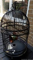 Photo showing entire black wrought iron bird cage with hanging decorative fish inside, placed on a black table outdoors near brick wall.