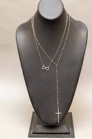 Two silver necklaces displayed on a black jewelry bust. One necklace has an infinity symbol pendant and the other has a cross pendant.