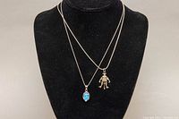 Two silver necklaces on black display; left pendant oval blue opal-like stone, right pendant articulated figure.