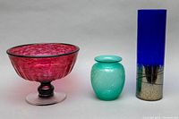Front angle shot showing the red-purple Ian Forbes pedestal bowl, small green frosted etched vase with lid, and tall cobalt blue cylindrical vase with silver effect base.