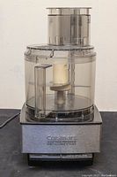 Front view of assembled processor showing stainless steel base, clear plastic work bowl with lid and feed tube