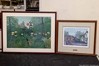 Two framed art prints side by side on a black surface, one larger left with jungle print, right smaller with street scene.
