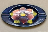 Top view of the Moorcroft porcelain ashtray showing the raised flower design with pink petals and green-yellow leaves against a dark blue background. Two cigarette rests are visible.