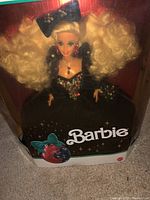 Close up of Barbie doll in original box, showing blonde curly hair, black dress with sequin embellishments, jewelry and original packaging.