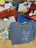 Eight men's XL shirts and sweatshirts laid out on a surface featuring Disney-themed prints, including Mickey Mouse and other Disney characters, in various colors.