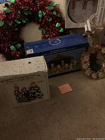 Photo of Christmas decorations including the nativity scene box, lighted musical Christmas tree box, red and green tinsel wreath, gold and white wreath, and candle ornament.