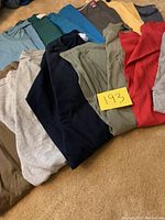 Twelve folded men's XL T-shirts in various colors including beige, blue, green, black, red, and gray, laid flat on carpeted floor.