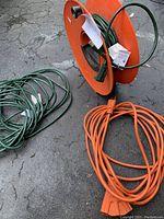 Three outdoor extension cords: a green one on orange holder, a loose green cord, an orange cord with triple outlet end lying on concrete floor.