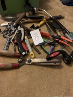 Photo showing a collection of garden tools laid on the floor, including hand spades, pruning shears, trimmers, and a hand saw in working condition as indicated by the seller.