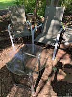 Two outdoor sling chairs with metal frames and grey mesh fabric next to two round glass-top side tables with metal legs outdoors in a shaded garden area.