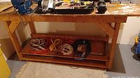 Wood workbench with mounted metal vise on left, showing front view with storage shelf underneath containing coiled cables (not included).
