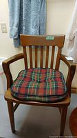 Front view of a wooden chair with armrests and vertically slatted back. Cushion with red and green plaid pattern on the seat.