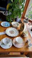 Photo showing various china pieces including Bunnykins plate, floral plates, tea cups, a small pitcher, egg holders, and decorative bowl arranged on a wooden table
