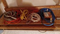 Photo showing multiple coiled electrical extension cords in various colors and lengths arranged on a wooden shelf.