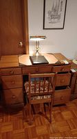 Wood desk with seven drawers, wooden chair with floral cushion, and brass desk lamp with dual white glass shades, positioned in a room with parquet flooring.