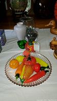 Clear glass plate holding a variety of colorful glass fruits and vegetables in realistic shapes and sizes, with a white vase and glass jar with marbles in the background.