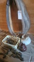 Side view of worn leather horse harness collar leaning against wall, basket of vintage keys in front, brown ceramic insulator and white ceramic lamp base on floor.
