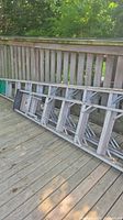 Two long silver folding aluminum ladders leaning horizontally against a wooden railing on a deck, showing full length and structure.