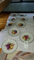 Nine round porcelain fruit plates arranged on a white cloth on wood floor, each with colorful fruit designs and gold trim