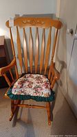 Full front view of vintage wooden rocking chair with floral cushion, showing carved floral detail on backrest and arm scratch marks