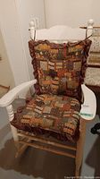 White wooden rocking chair with spindle arms and legs, featuring patchwork quilt patterned cushions with burgundy ruffled borders. Visible scratches on wood surface.