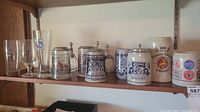 Full shelf view displaying five ceramic beer steins with lids including commemorative pieces and three clear drinking glasses.
