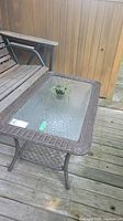 Outdoor wicker coffee table with clear textured glass top and lower wicker shelf, positioned on wooden deck with adjacent outdoor seating bench.