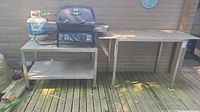 Both stainless steel tables shown side by side outdoors on a wooden deck, one with a propane grill and tank on top.