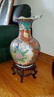 Full view of large colorful ceramic vase on wooden stand showing floral and bird details with scalloped rim.